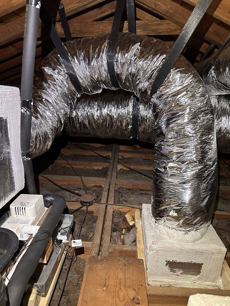 Insulated silver HVAC ducting in an attic with exposed wooden beams and a vent box attached to a concrete support
