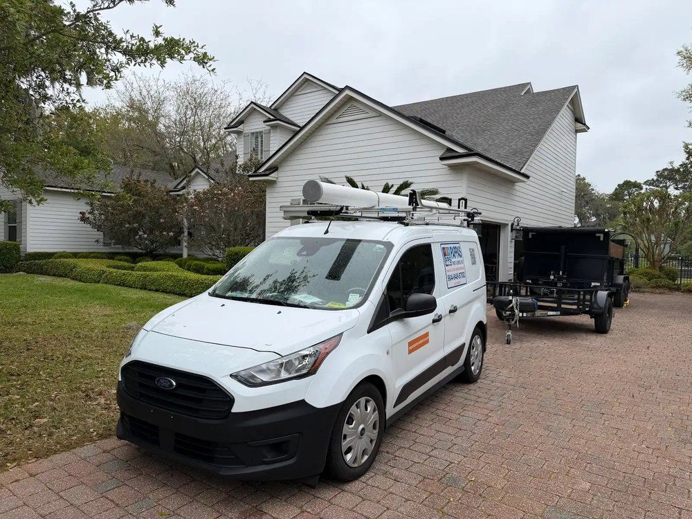 White service van with ladder rack parked in a driveway beside a black trailer near a house