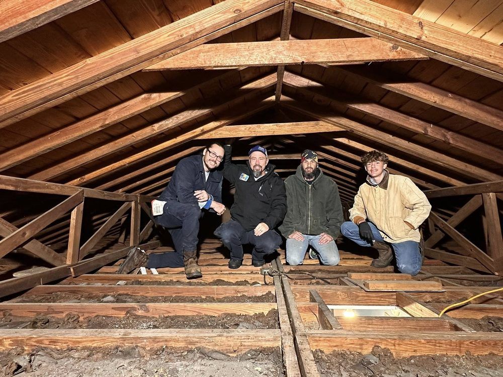 Four people sitting in an attic with exposed wooden beams and insulation strips under construction.