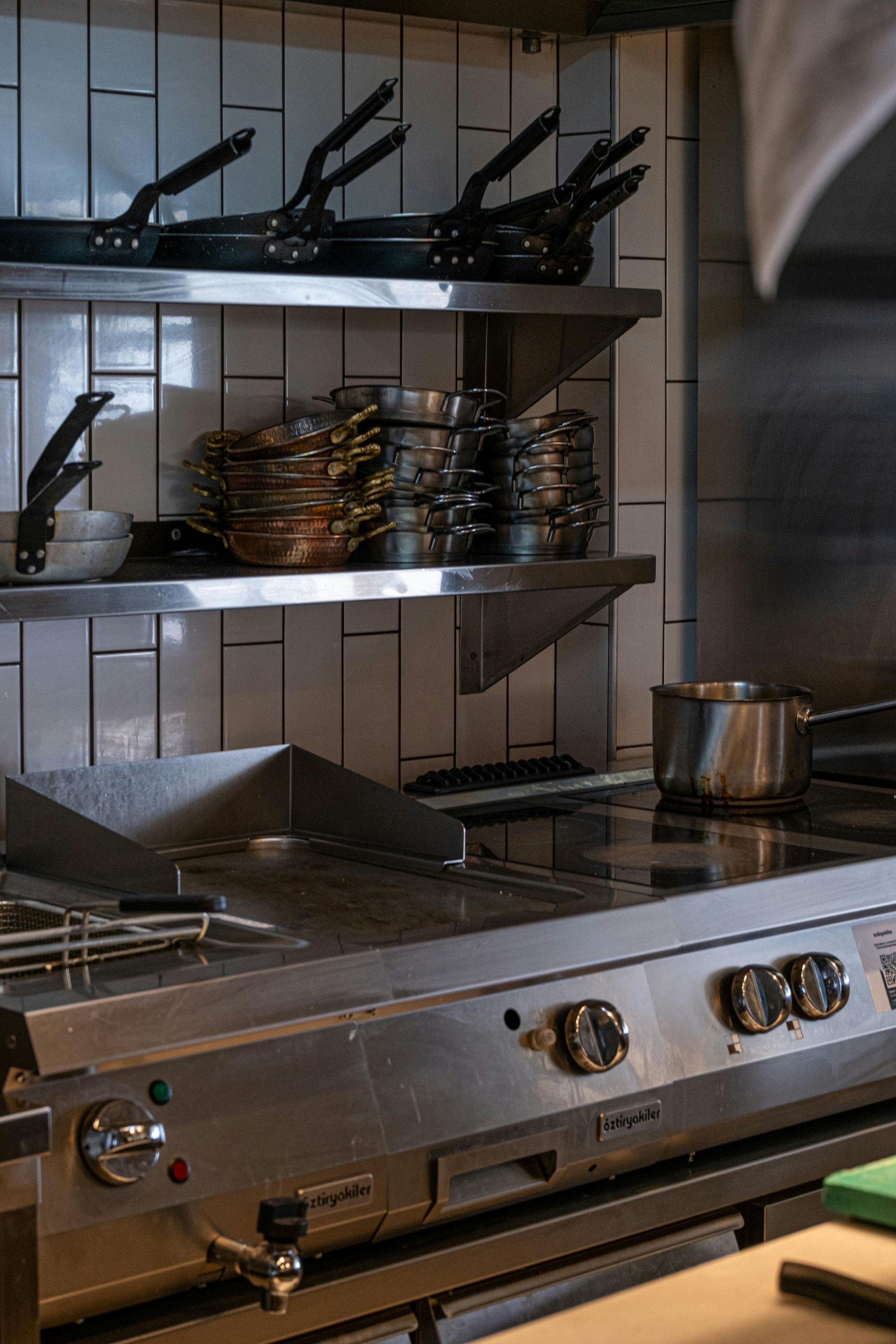 Commercial stainless-steel kitchen stove with pans, pots, and utensils on shelves above the counter.