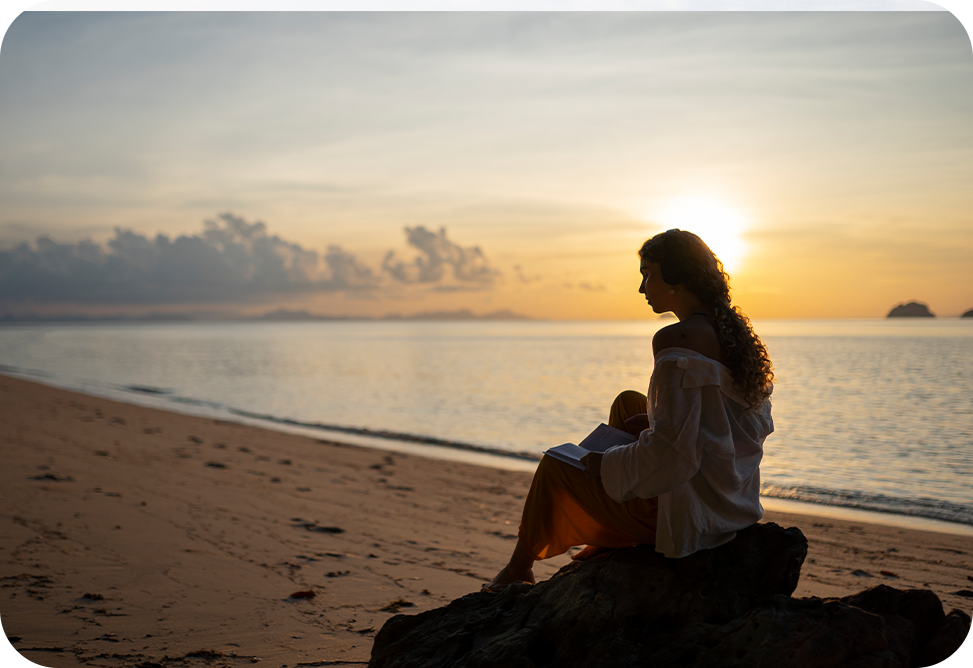 A woman is sitting on a rock on the beach at sunset.