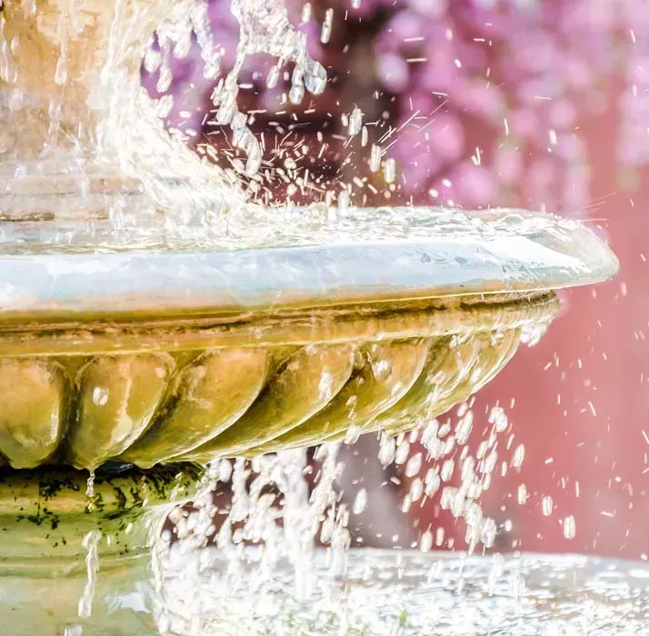 Water fountain, close-up view; water cascades over the ornate, aged stone structure, with pink, blurred background.