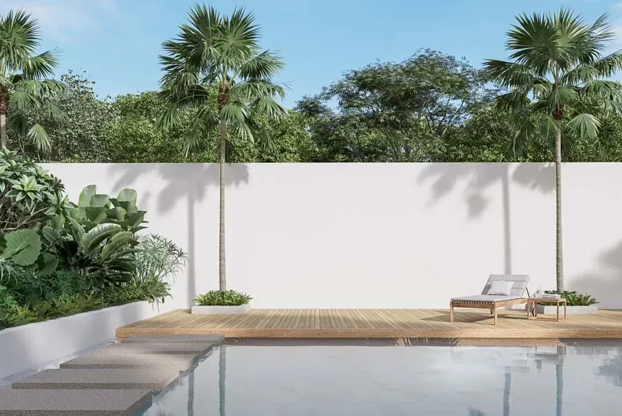 Poolside scene with a white wall, deck, lounge chair, palm trees, and lush greenery under a blue sky.