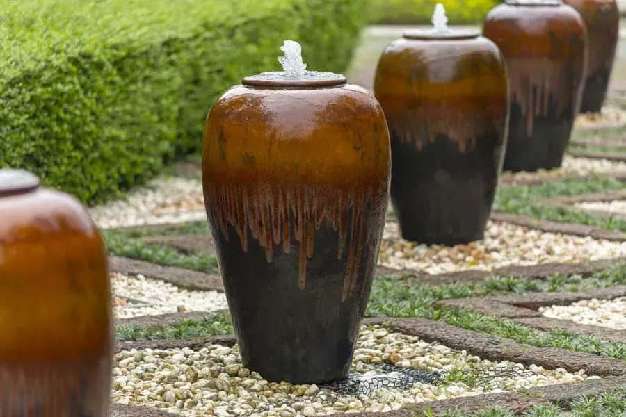 Row of glazed brown water fountain urns in a landscaped garden, with water spouts.