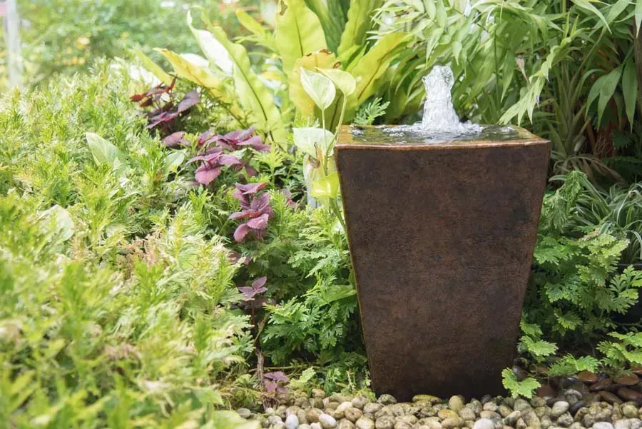 Water fountain in a garden setting, with plants and pebbles. Brown, tapered fountain sprays water.