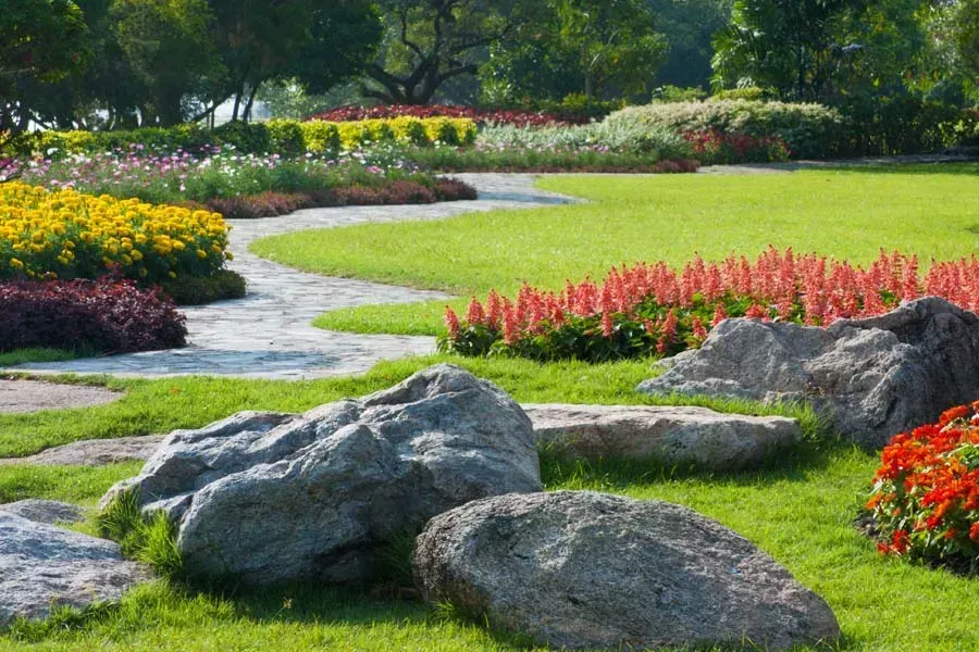 Green lawn with winding stone path, colorful flowerbeds, and large rocks.