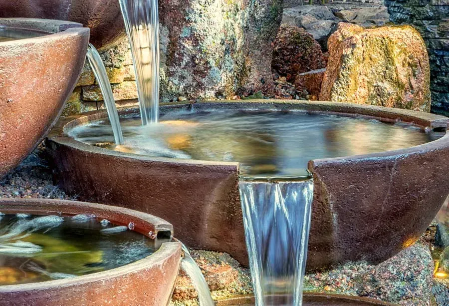 Water cascading from brown, oval-shaped ceramic bowls in an outdoor fountain against a rocky backdrop.