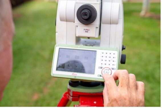 A person uses a total station surveying instrument mounted on a tripod outdoors, measuring.