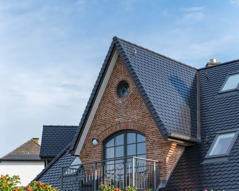 Core One Construction | Brick and dark tile roof with dormer, arched window, and small balcony against a blue sky.
