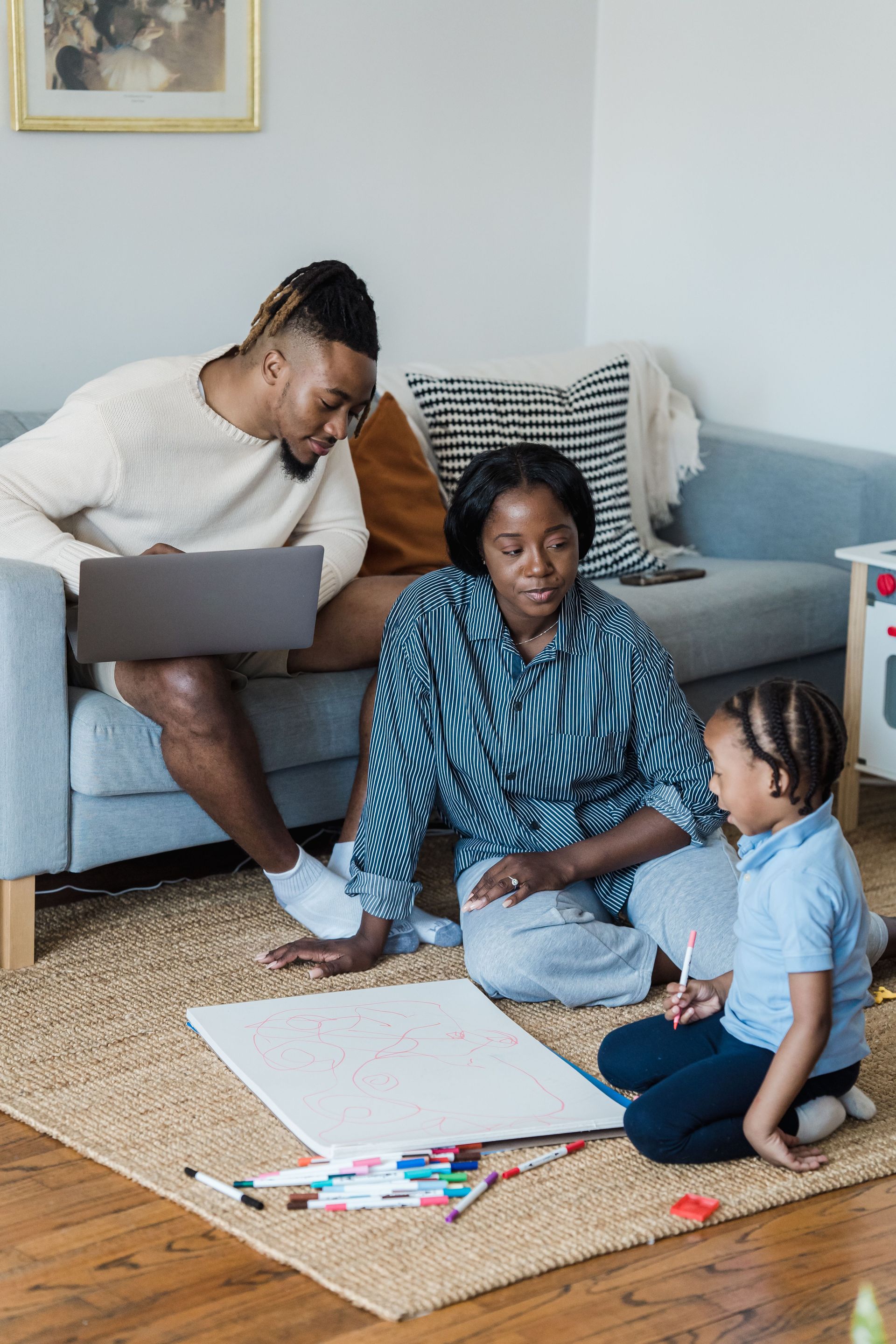 Family drawing together on the living room floor while one adult uses a laptop on the couch.
