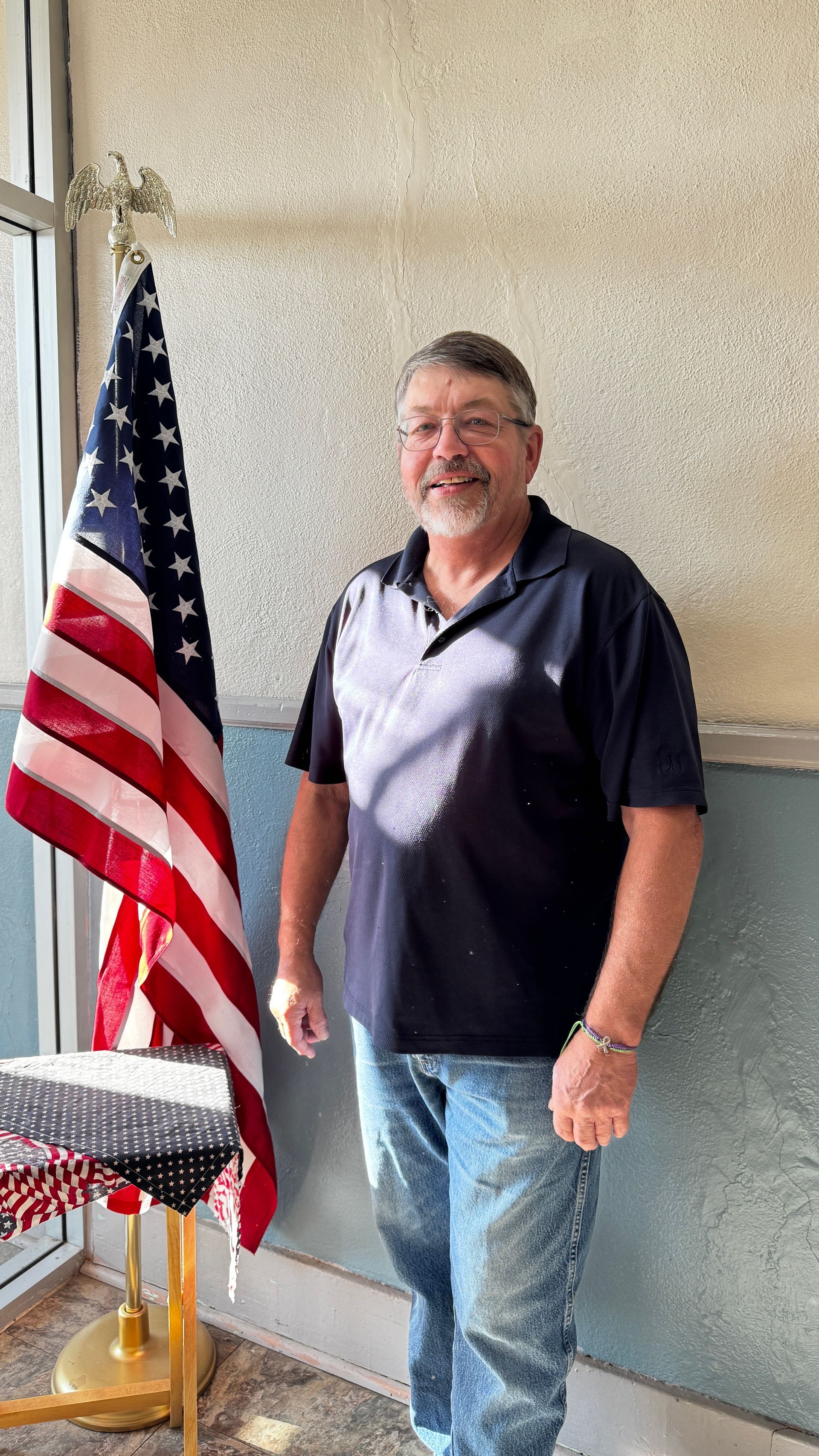 Man in front of an American flag smiles. He wears a blue shirt, jeans, and glasses.