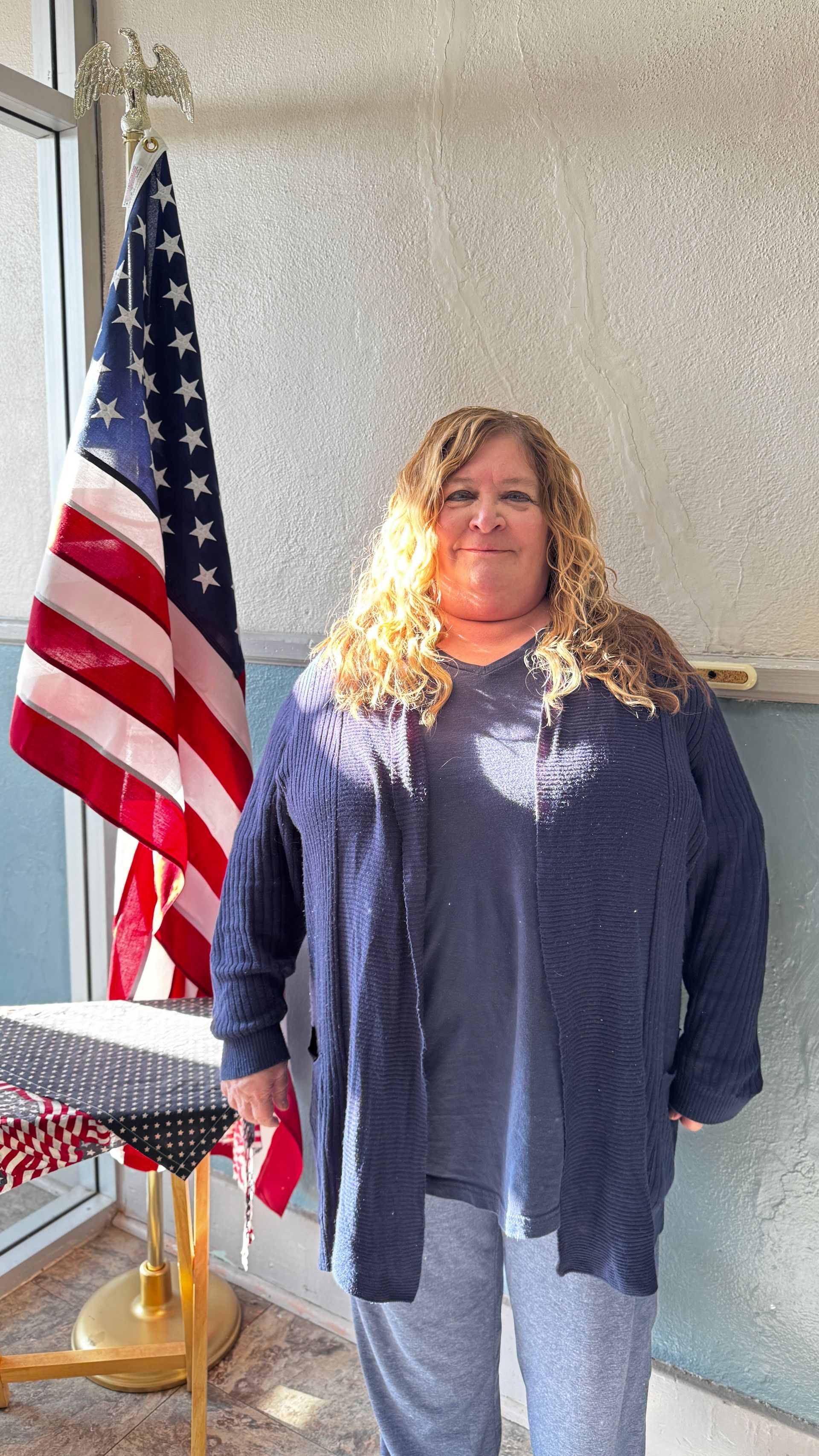 Woman in blue sweater stands beside an American flag in front of a window.