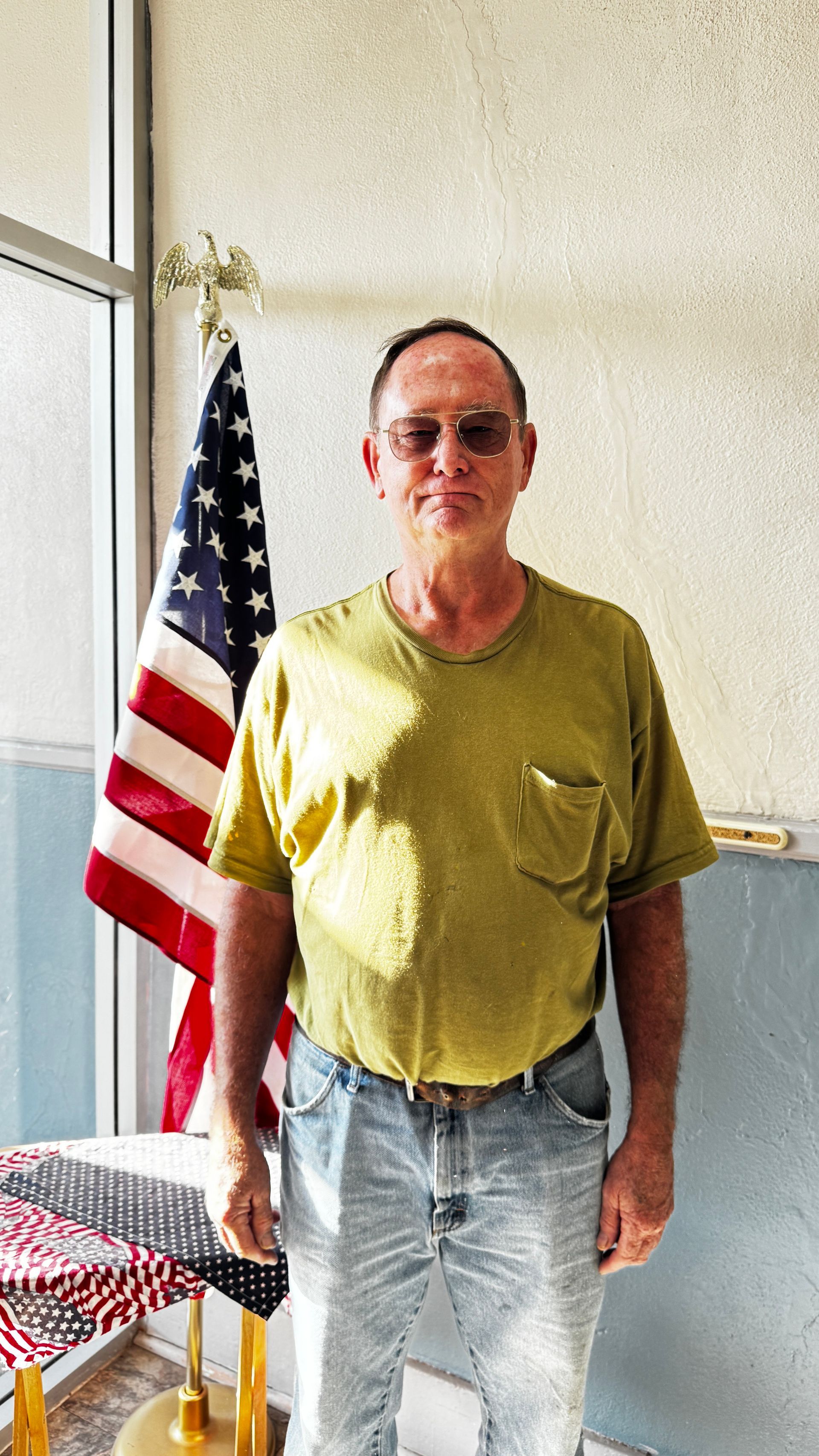 Man in yellow shirt and jeans stands in front of an American flag.