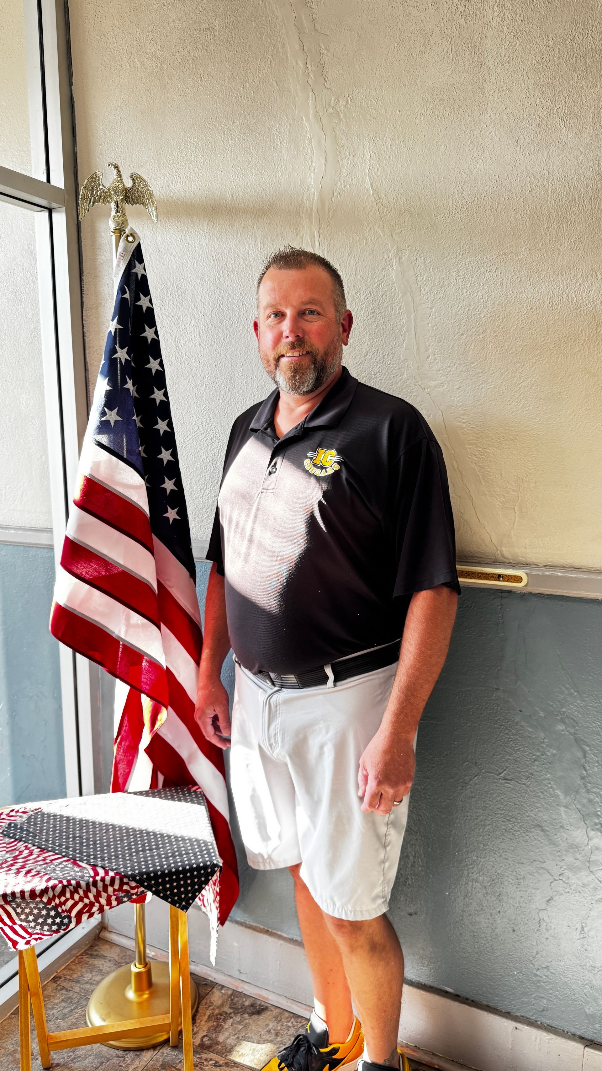 Man stands next to American flag. He wears a black polo and white shorts.