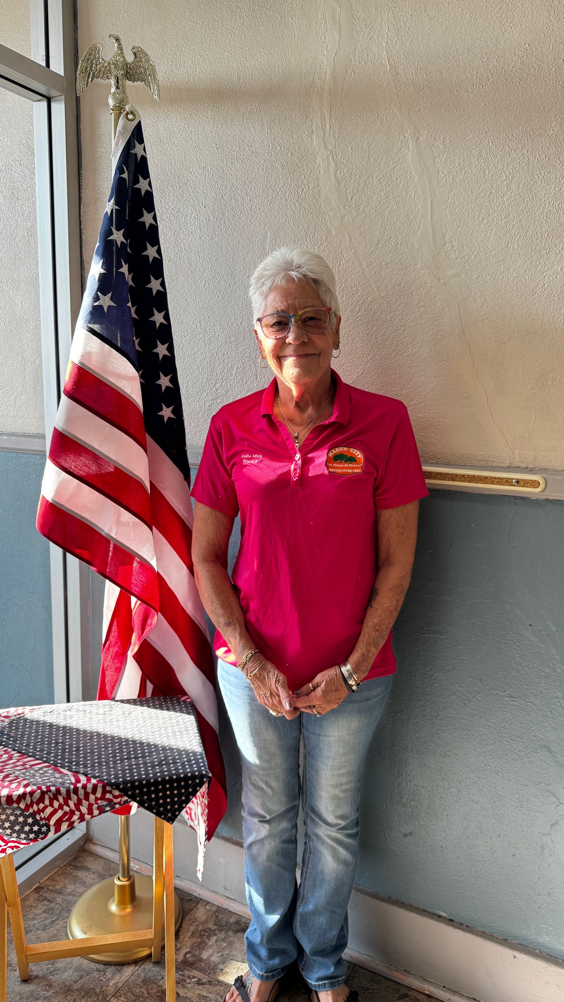 Woman with short gray hair in pink shirt and jeans stands by US flag.
