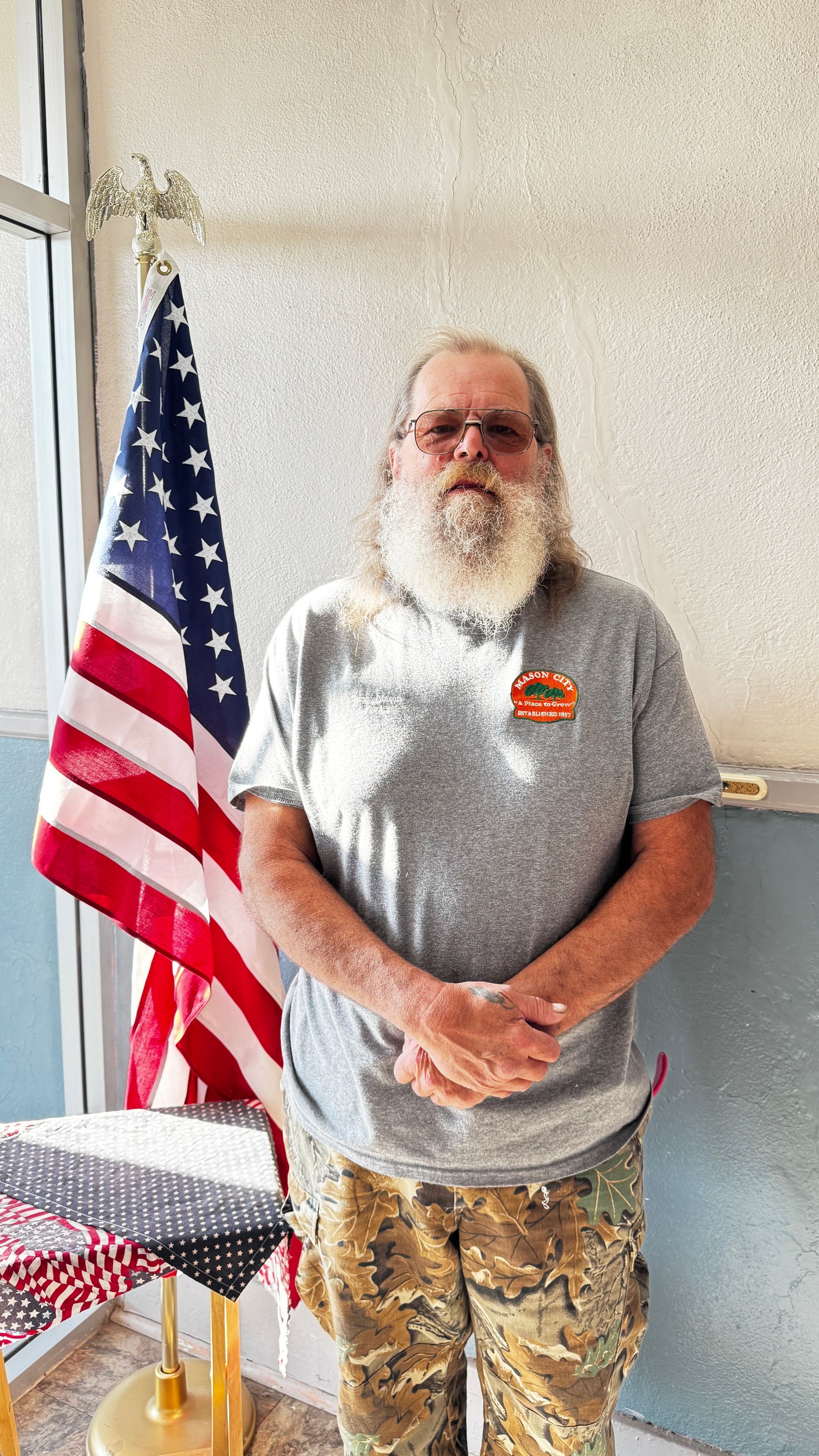 Man with beard and camouflage pants stands in front of an American flag.
