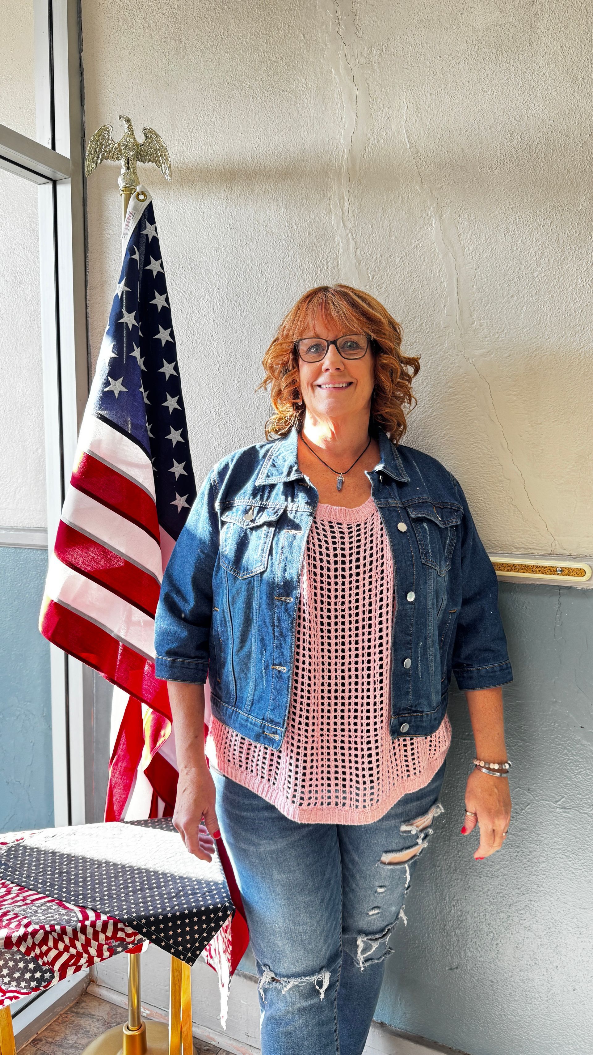 Woman in denim jacket and jeans stands near an American flag indoors, smiling.