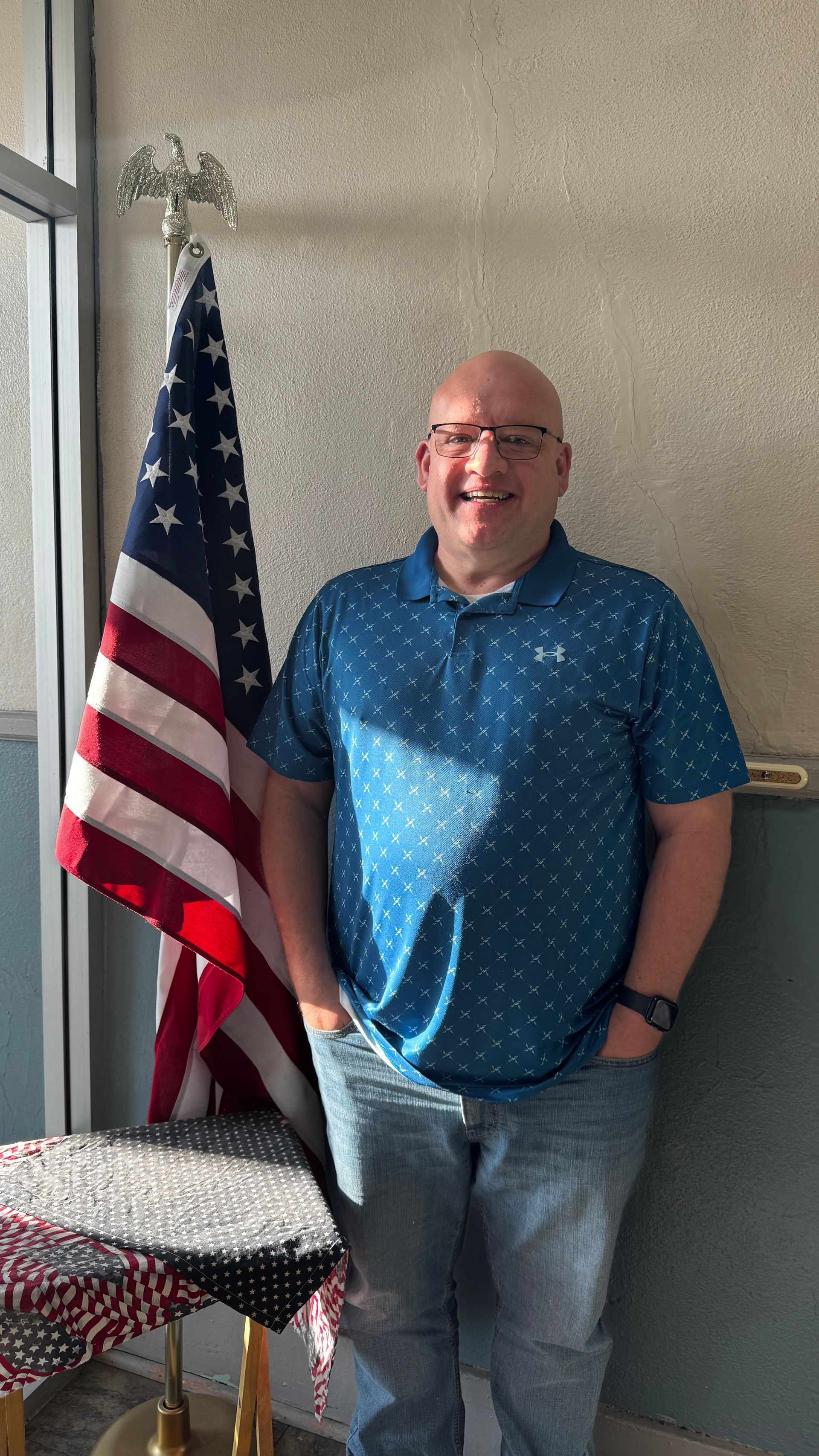 Man standing next to American flag, smiling, wearing glasses, blue patterned shirt, jeans, and leaning against a wall.