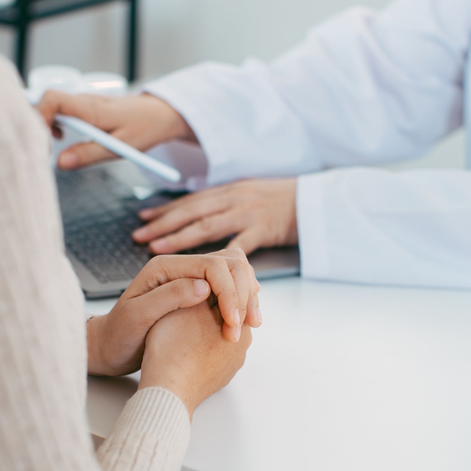 A person sits with folded hands while a healthcare provider in a lab coat types on a laptop during a consultation.