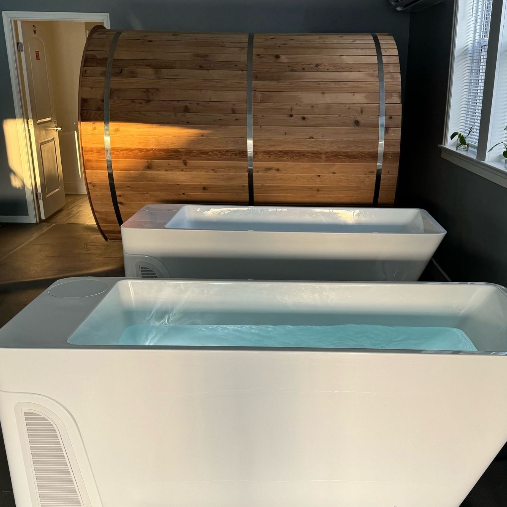 Two white, rectangular cold plunge tubs sit in a room with a wood-paneled sauna in the background.