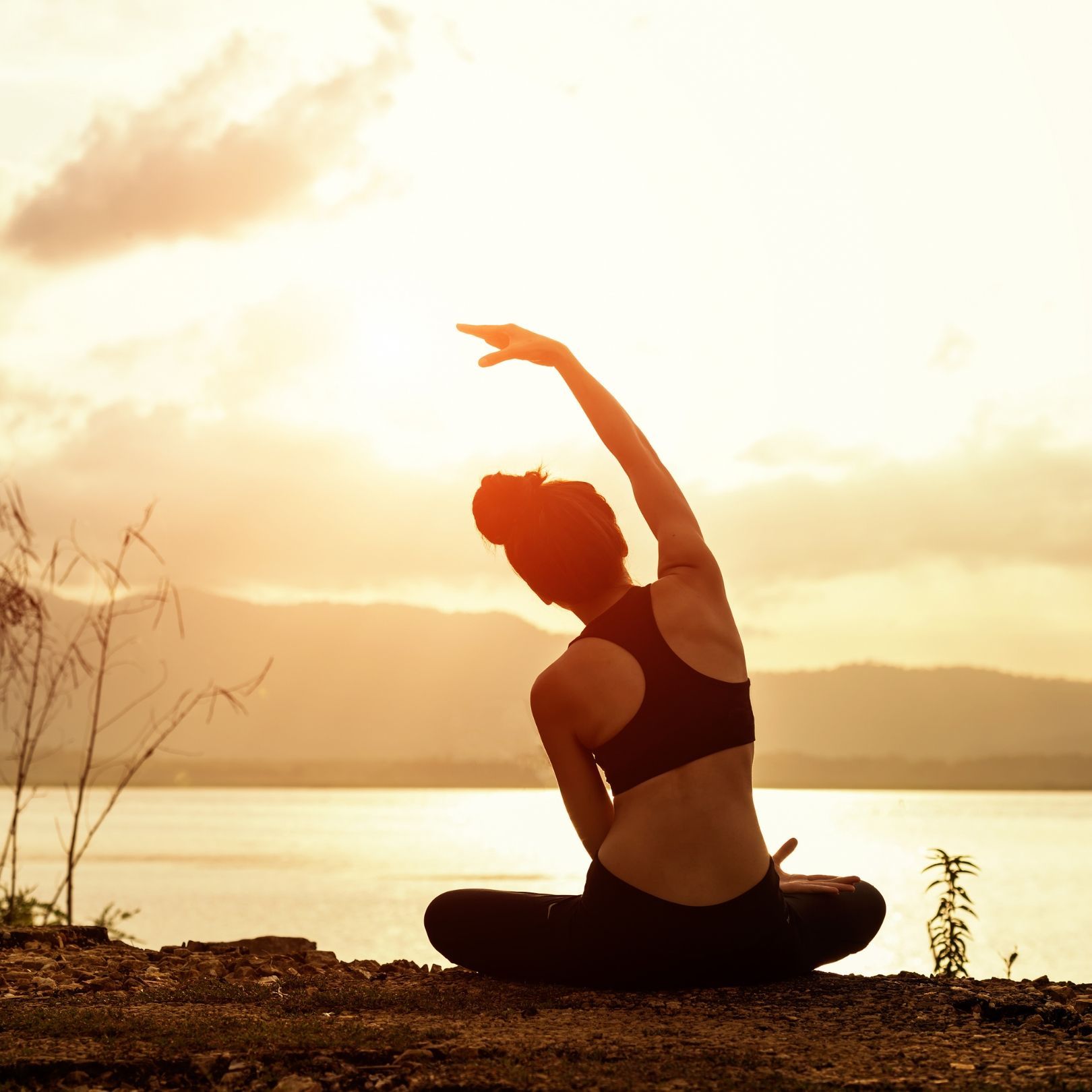 A silhouette of a person practicing yoga in a seated side stretch by a lake during a golden sunset.