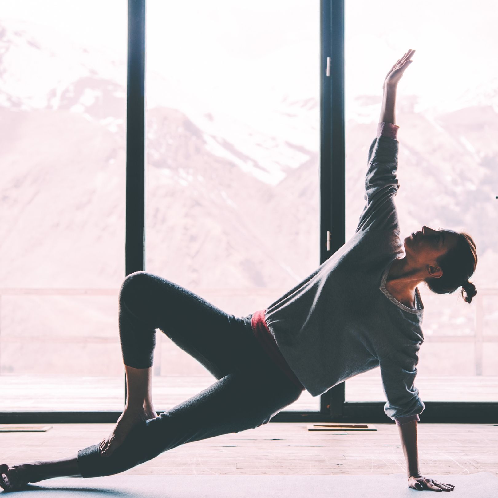 A person in athletic wear performs a side plank yoga pose with one leg lifted, against a window with a mountain view.