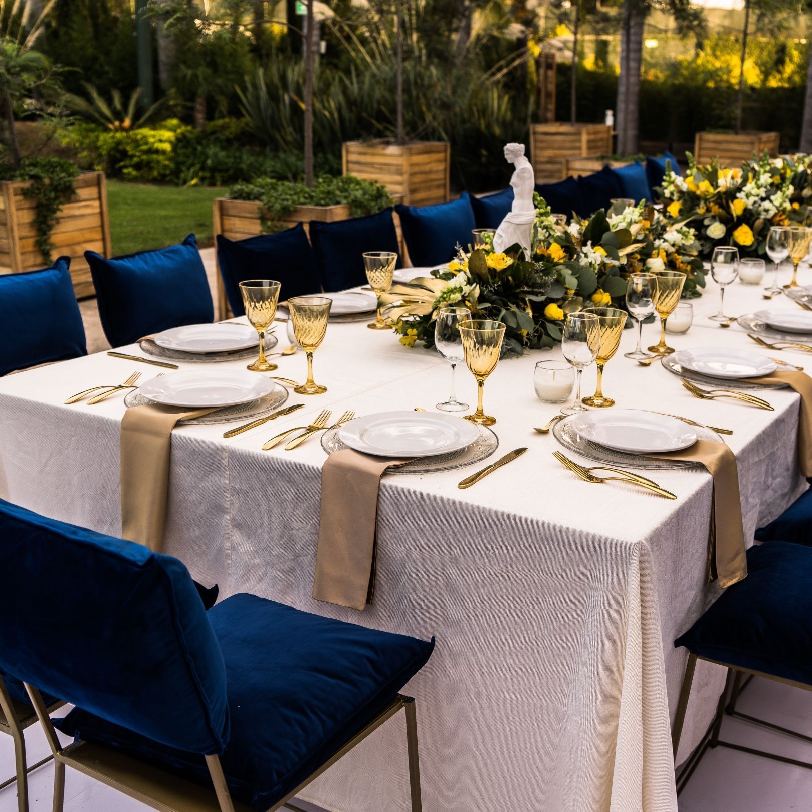 An elegantly set outdoor dining table with white linens, gold accents, blue velvet chairs, and floral centerpieces.