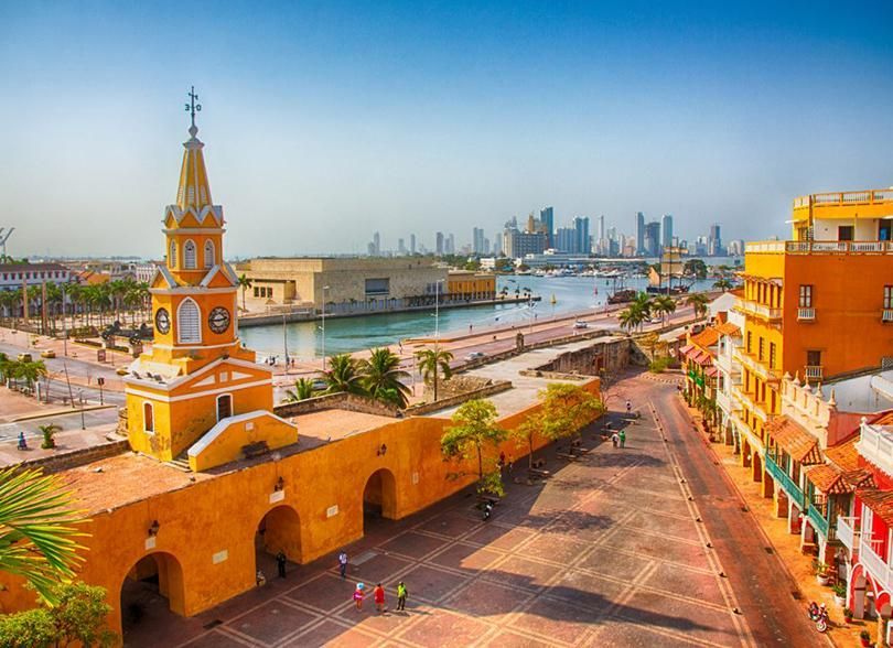 Paisaje urbano ribereño lleno de color, con la torre de una iglesia, edificios de color naranja y un horizonte lejano al atardecer.