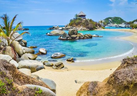 Playa tropical soleada con agua turquesa, rocas, palmeras y una pequeña cabaña isleña en la distancia.