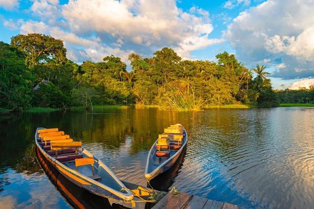 Dos barcos de madera en un río tranquilo al atardecer, con frondosos árboles verdes y un cielo nublado reflejado en el agua.