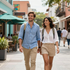 Dos personas caminando y sonriendo en una soleada calle comercial al aire libre, con cafés y tiendas cerca.