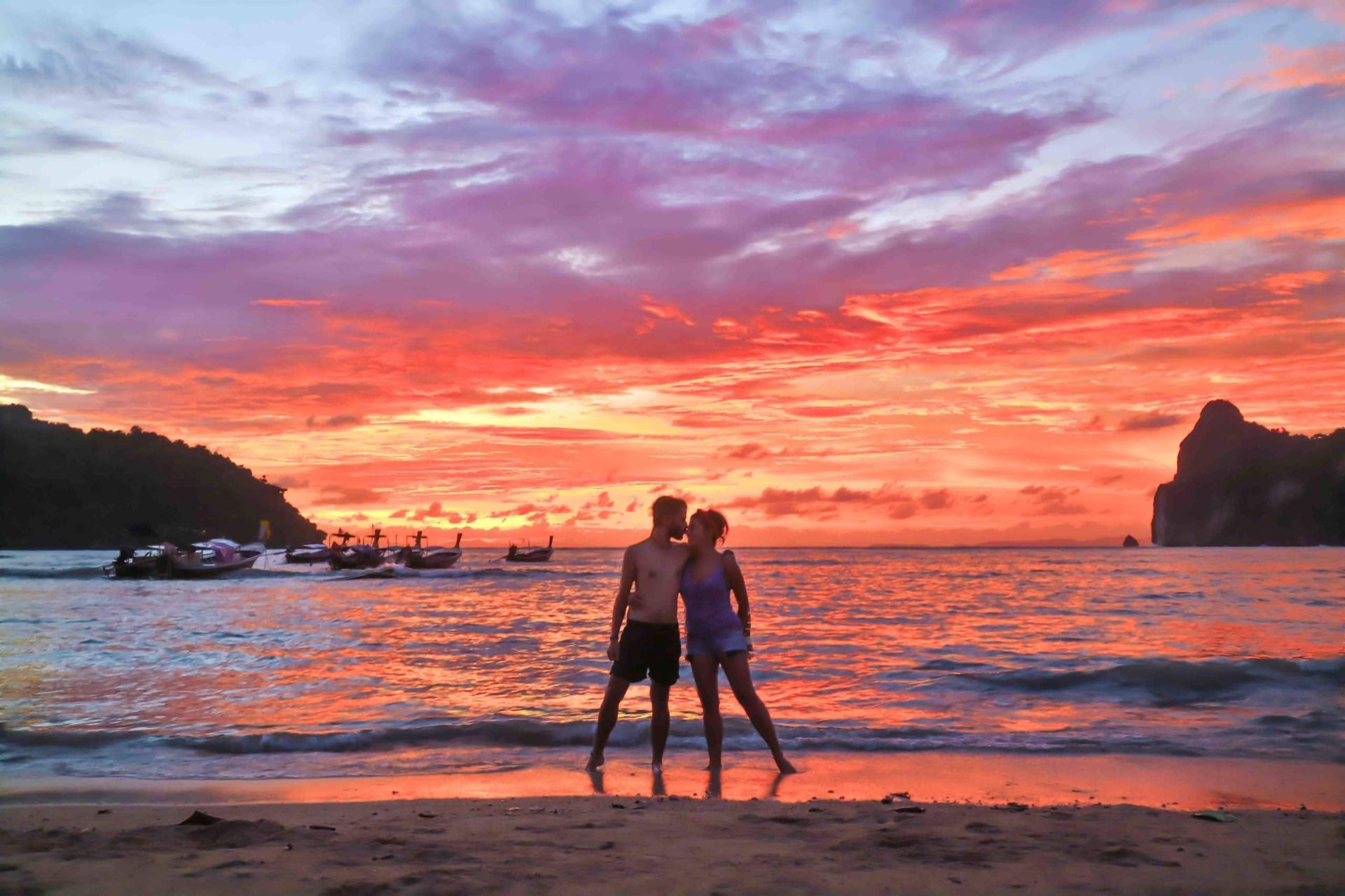 Dos personas de pie en una playa al atardecer con barcos en el agua y cielo rosa anaranjado.