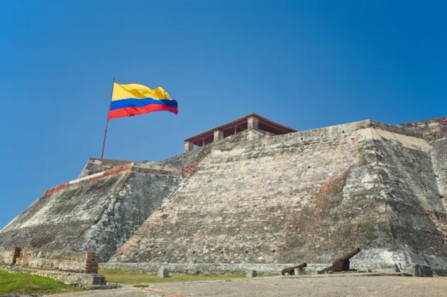 Gran fortaleza de piedra con una bandera amarilla, azul y roja ondeando contra un cielo azul despejado.
