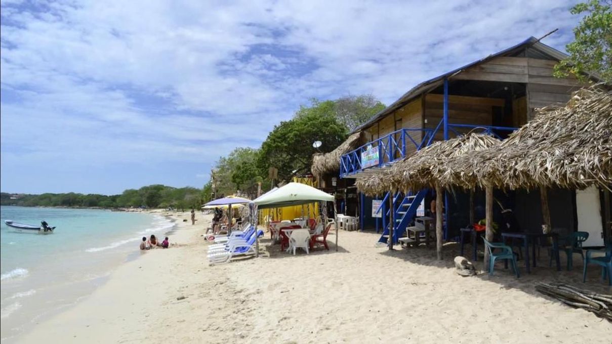 Playa soleada de arena blanca, sombrillas de paja, tumbonas y una cabaña junto al mar, al lado de aguas cristalinas.