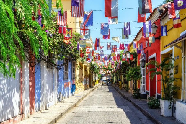 Calle estrecha y colorida con banderas ondeando, plantas en macetas y edificios brillantes bajo un cielo soleado.