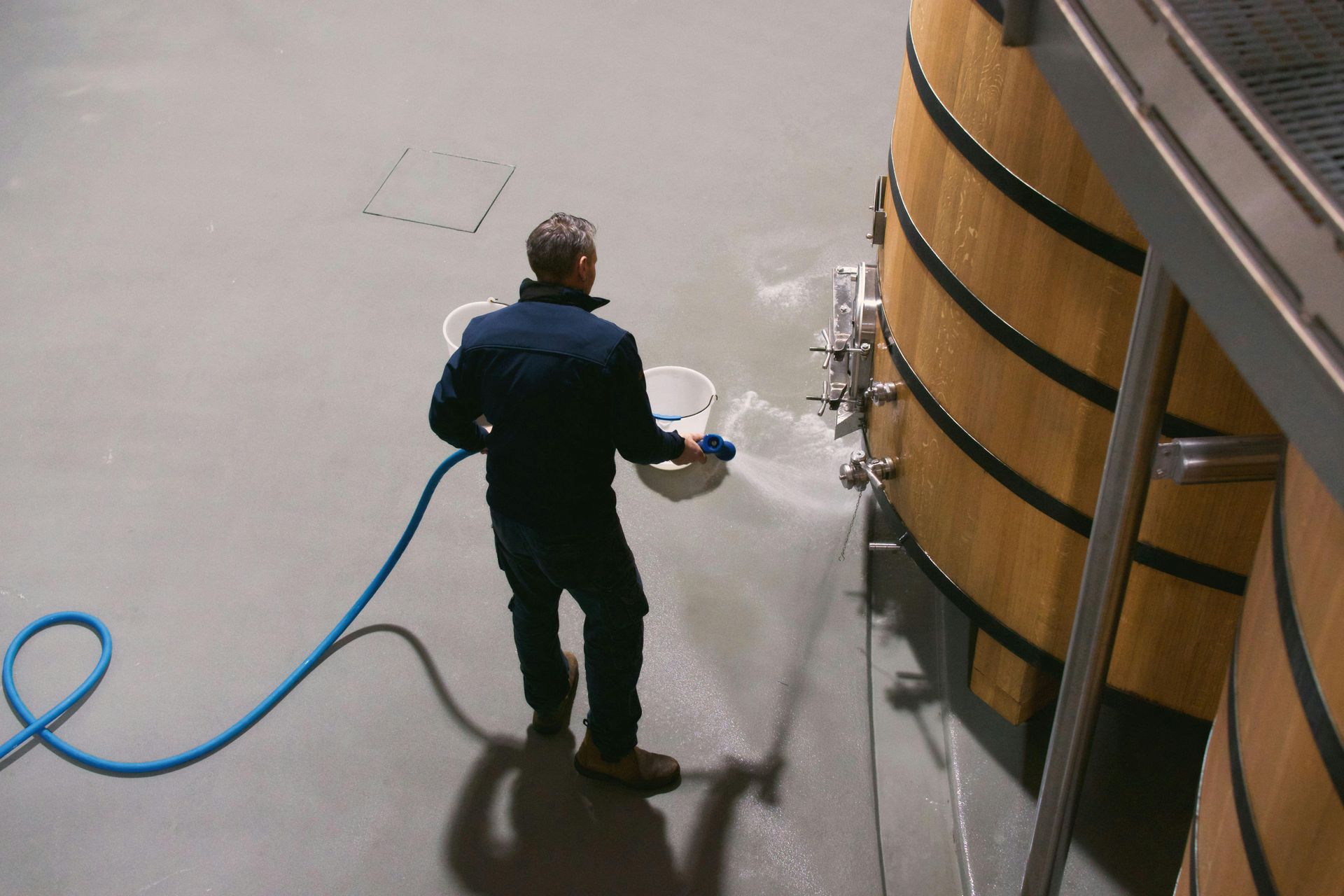 Man cleaning near a large wooden wine tank with a hose and buckets in a winery.