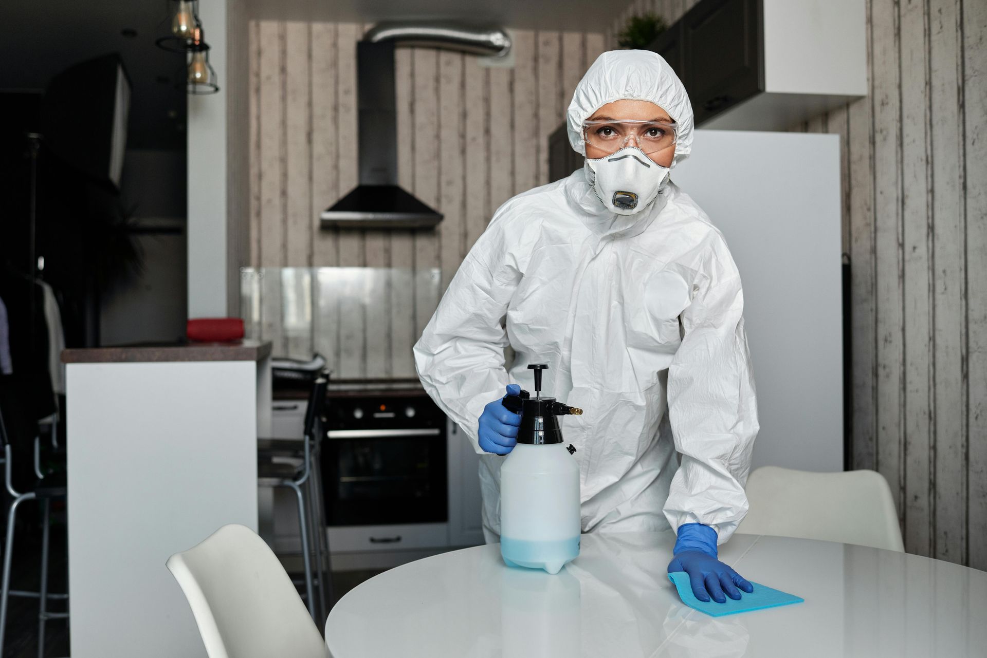 Person in hazmat suit disinfecting a kitchen table with a spray bottle and cloth.