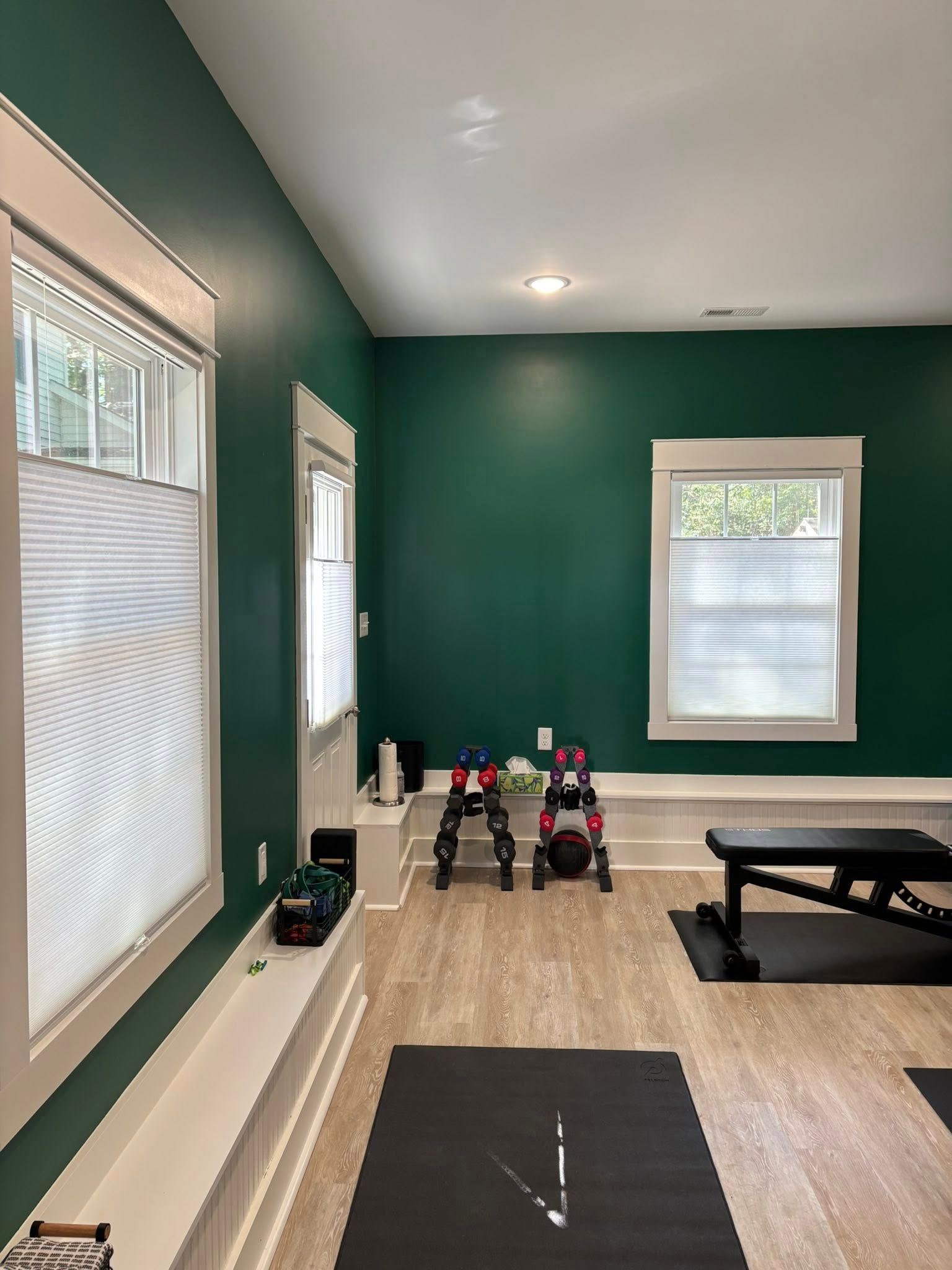 Home gym with green walls, windows, mat, dumbbells, and a small exercise bench on wood floors