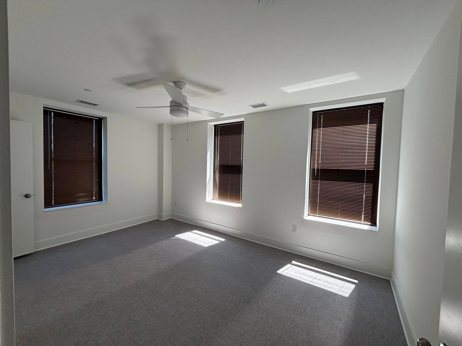 Empty white bedroom with gray carpet, ceiling fan, and three windows with blinds casting sunlight on the floor