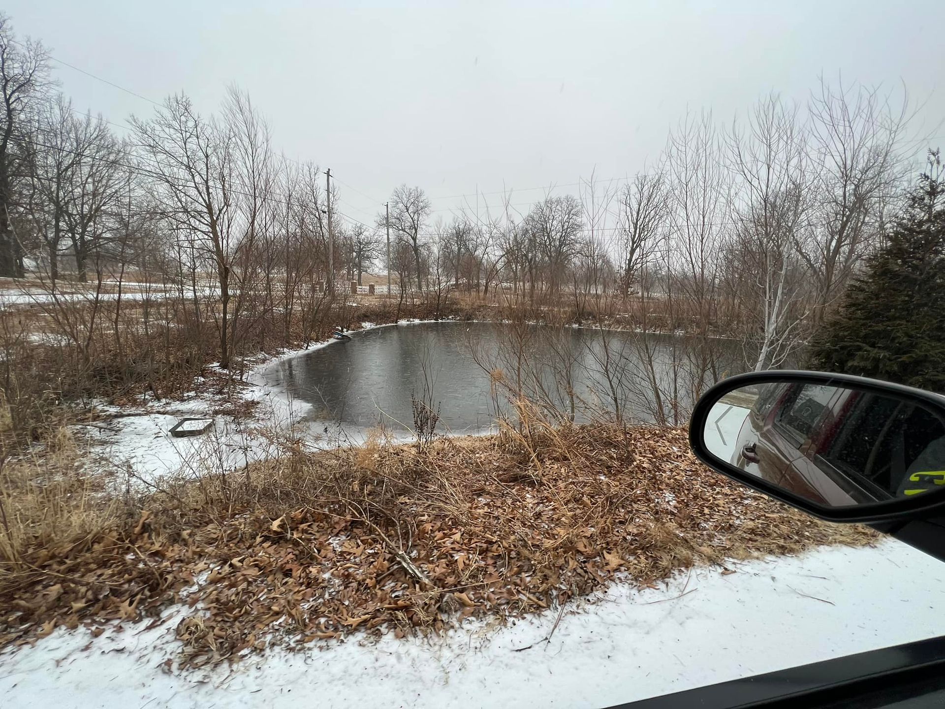 A pond surrounded by bare trees and snowy ground, seen from a car window on a cloudy day.