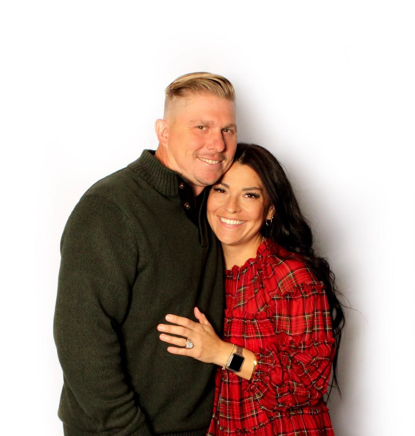 Couple smiling and embracing, man in green sweater, woman in red plaid dress, white background.