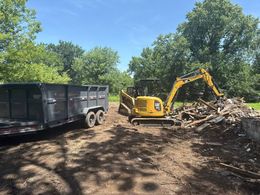 Yellow excavator loading debris into a trailer on a dirt lot under a blue sky with trees.