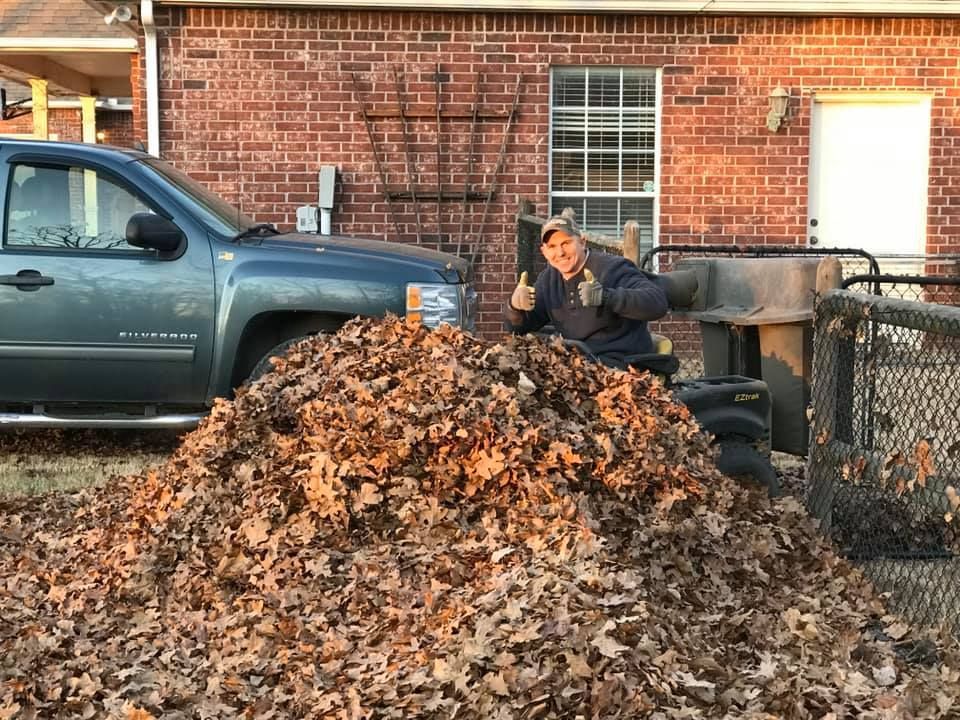 Man with thumbs up, posing by large pile of brown leaves, truck in background.