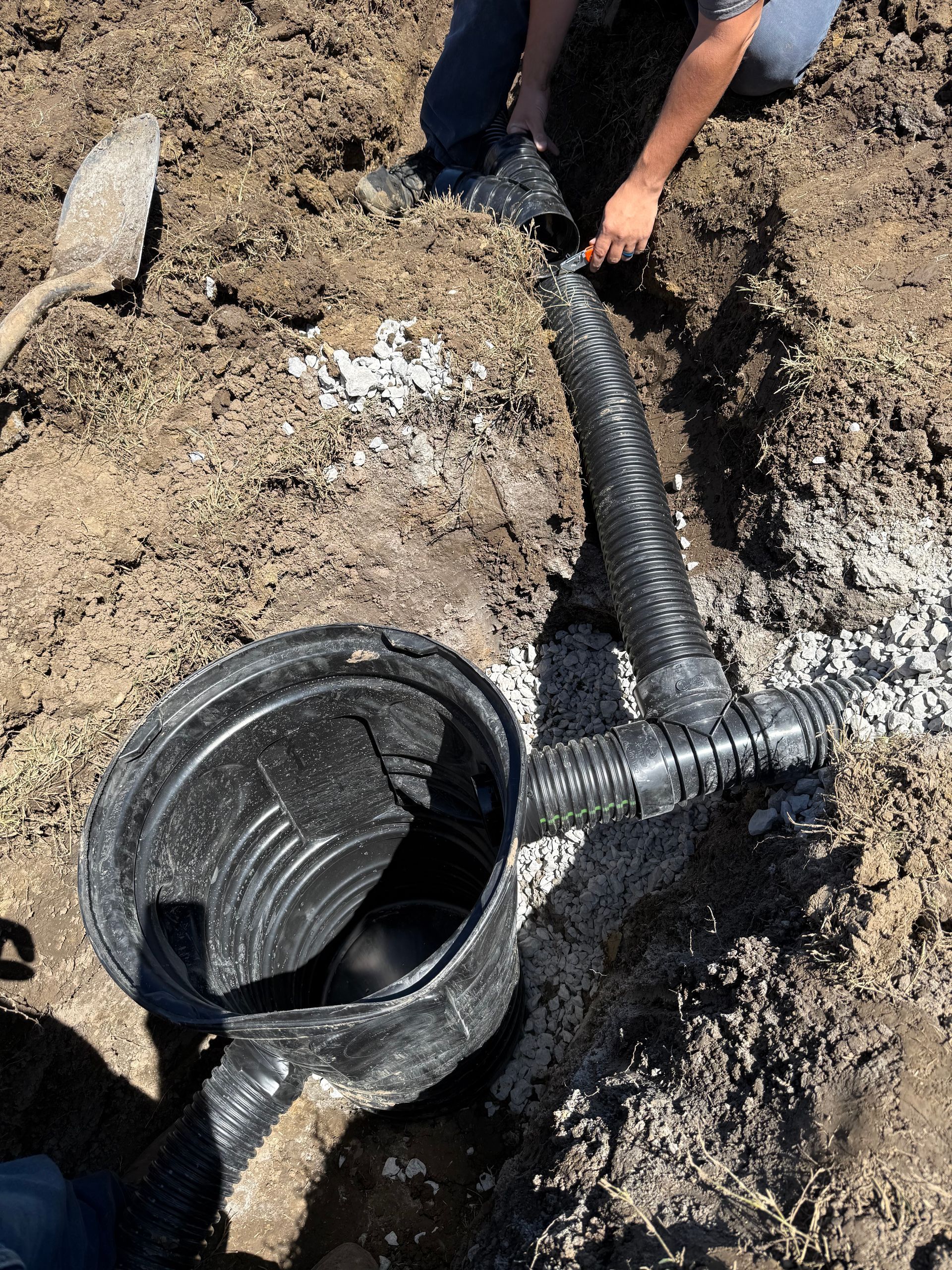 Two black corrugated pipes discharging water into a shallow stream, near dry grass.