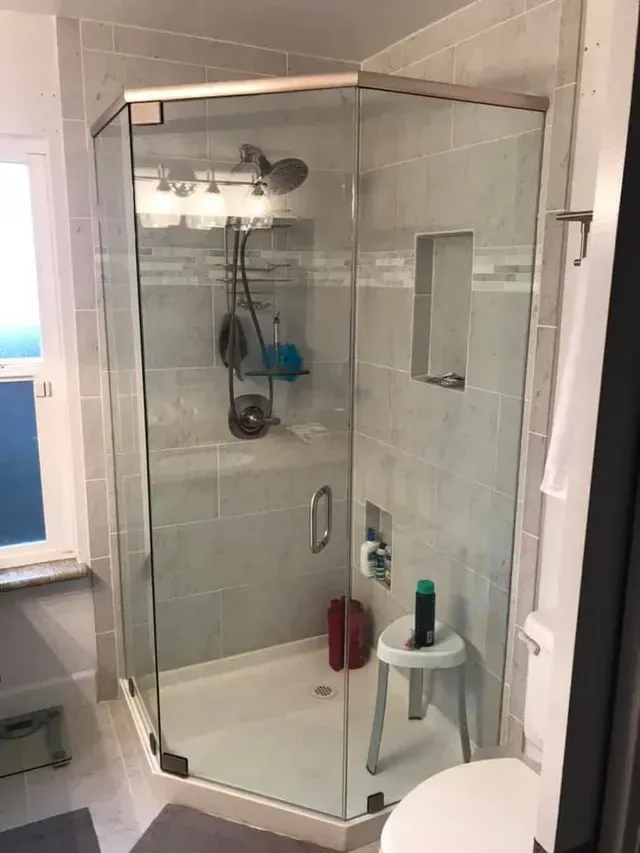 Corner shower with glass doors, gray tile, chrome fixtures, and a white stool.