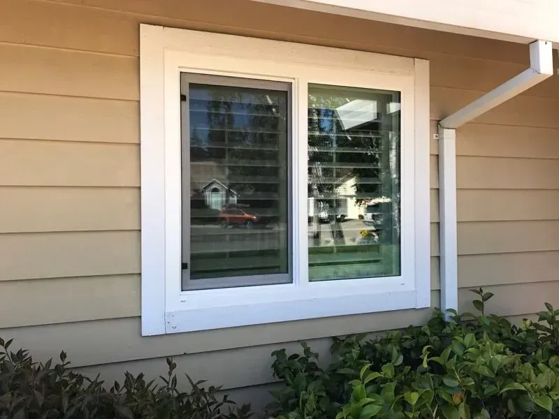 White-framed window on a tan house with a partially open window and greenery below.