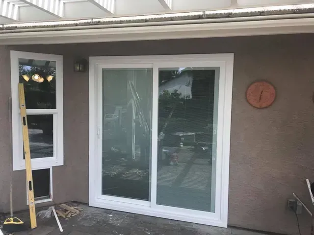 White-framed sliding glass door and adjacent window on brown stucco wall. A level leans against the window.