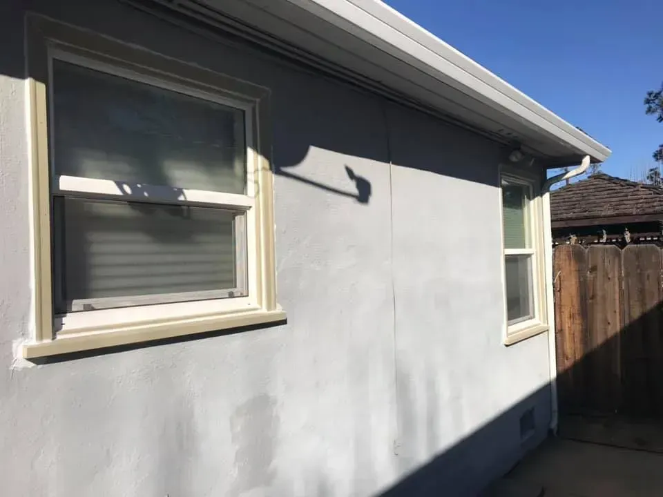Gray stucco wall with two windows, a white gutter, and a wooden fence. Sunny day.