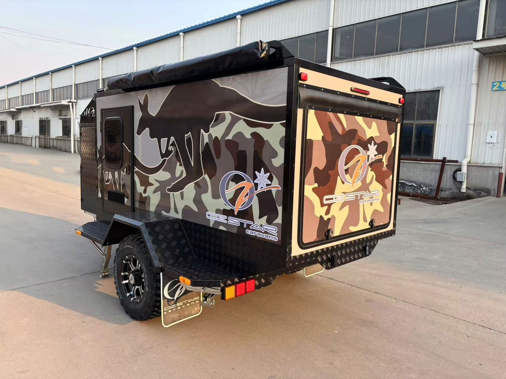A small 7ft caravan, trailer with a camouflage design is parked in front of a building.