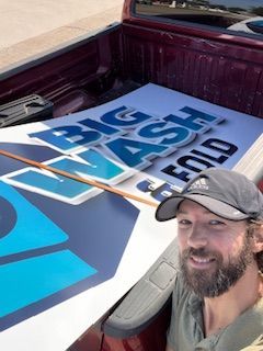 A man is sitting in the back of a truck holding a sign that says big ash fold